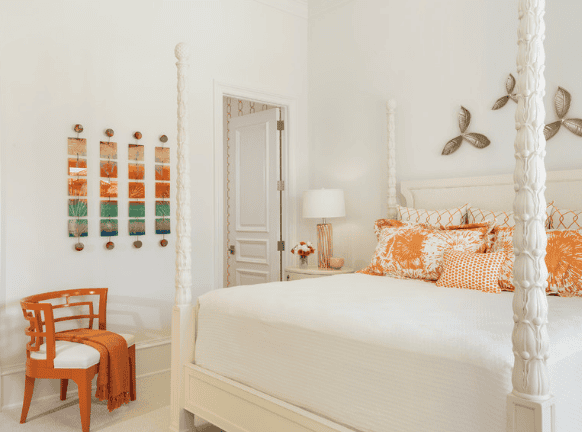 A bedroom with a white four-poster bed, orange and cream pillows, a white table lamp, a wooden chair with an orange blanket, and wall decor with leaf-shaped ornaments.