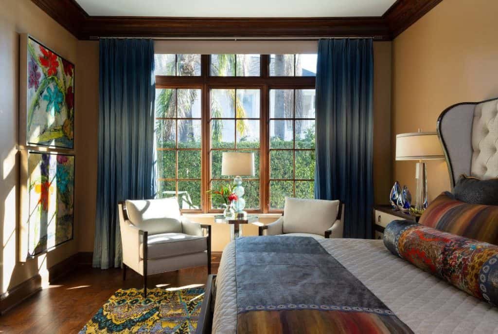Sunlit bedroom with large window, blue curtains, white armchairs, a side table with a lamp and flowers, colorful artwork, and a bed with patterned pillows.