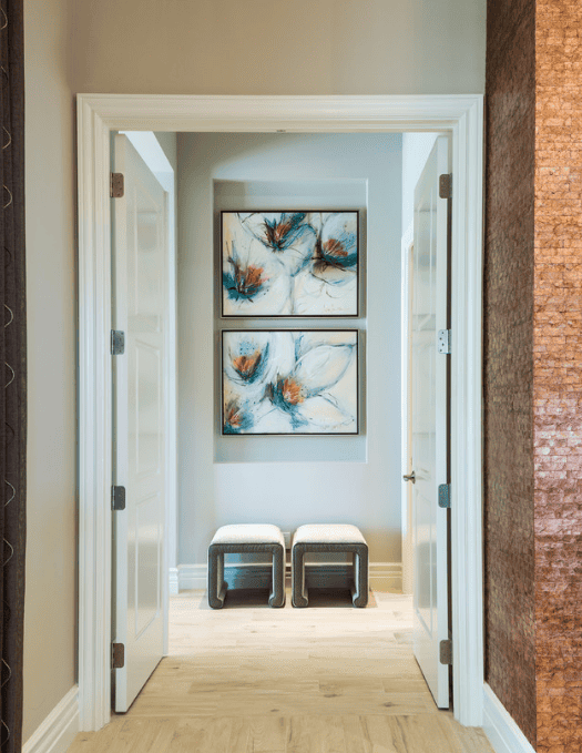 A hallway with open white double doors leading to a small sitting area with two stools and two abstract paintings on the wall.