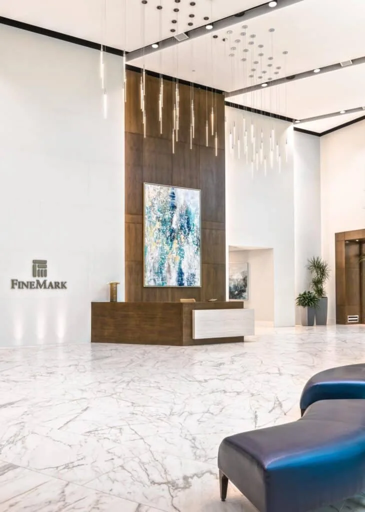 Modern corporate lobby with white marble floors, a wooden reception desk, abstract artwork on the wall, plants, and contemporary lighting fixtures on the ceiling.