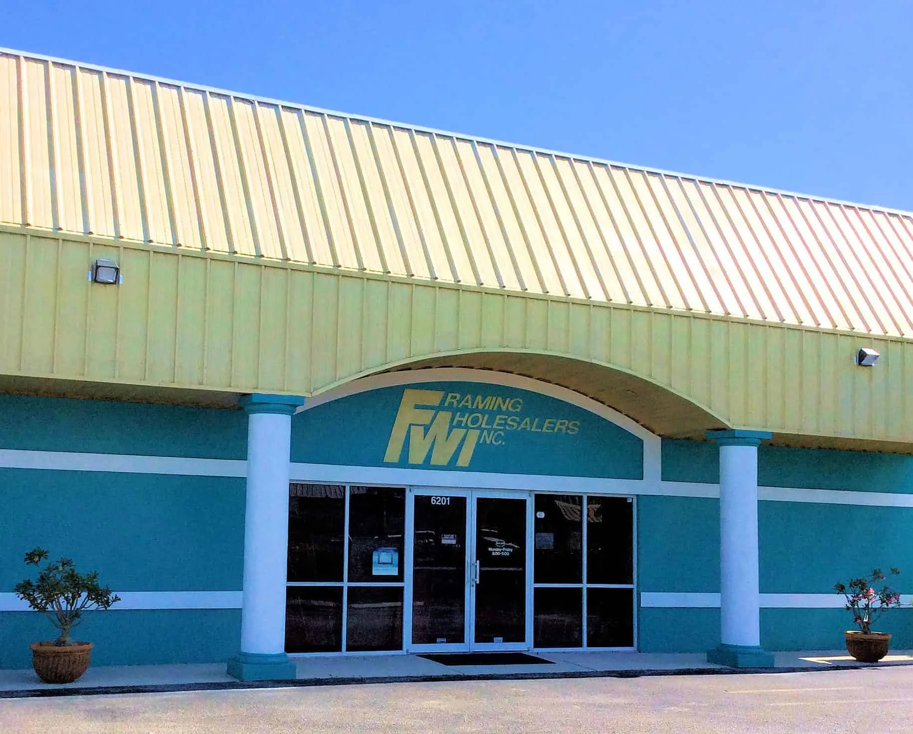 Front entrance of a building with teal walls, white columns, and a yellow roof overhang, featuring a sign that reads "Raming Holesalers Inc." The entrance has glass doors and two potted plants on each side.