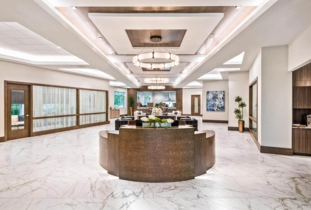 Luxury hotel lobby with white marble floors, modern lighting fixtures, and wooden accents. There are seating areas, artwork, and potted plants.