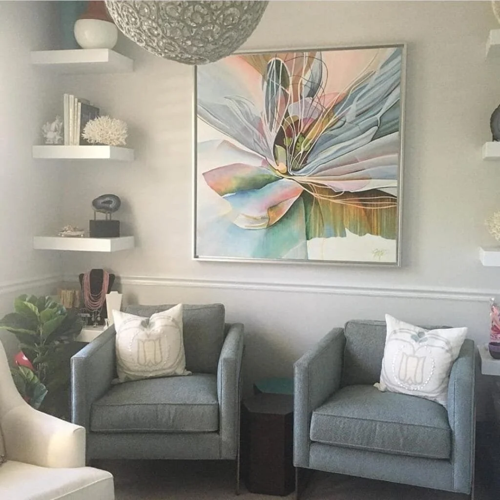 A living room corner with two gray armchairs, each with a white pillow, a small black side table between the chairs, a large colorful abstract flower painting on the wall, floating white shelves with decor items, and a large round pendant light.