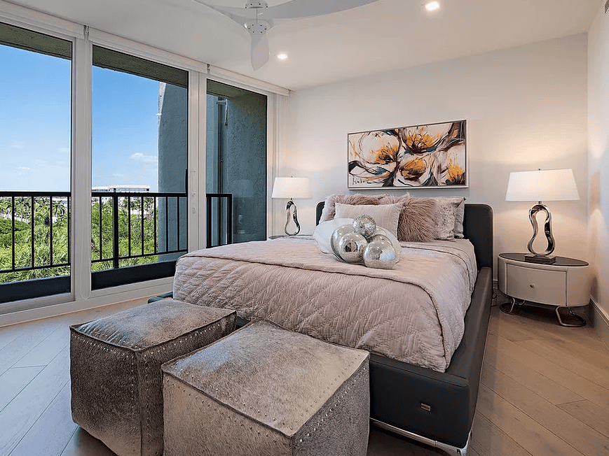 A modern bedroom with large sliding glass doors leading to a balcony, a bed with beige bedding and decorative pillows, two silver ottomans at the foot of the bed, a black headboard, two matching white lamps on nightstands, and wall art above the bed,