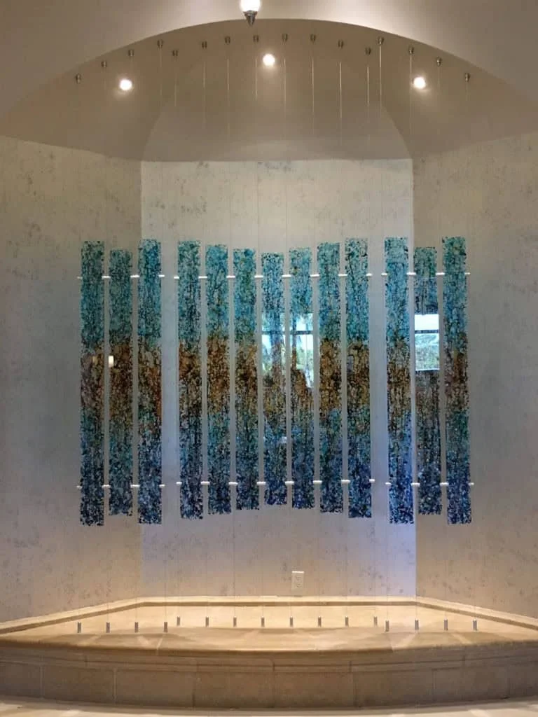 Interior of a room with a large artwork featuring blue, gold, and green colors, framed by vertical slats, with a window visible behind it and ceiling lights overhead.