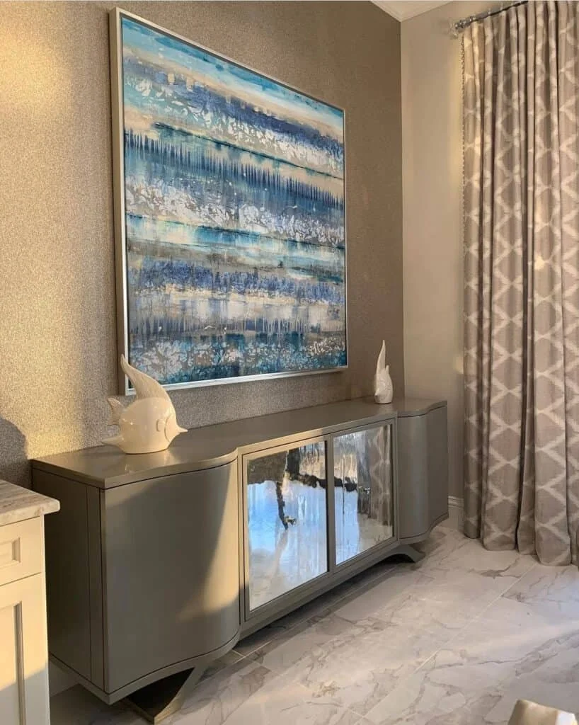 A modern sideboard with mirrored sliding doors, positioned against a textured wall. On top, there are two white decorative sculptures resembling abstract sea shells. Above the sideboard hangs a large abstract painting with blue, white, and beige hori