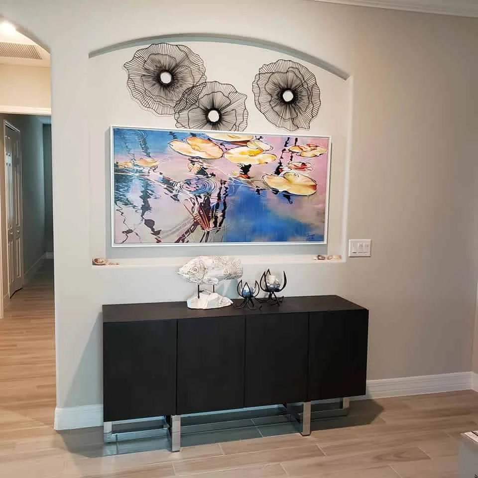 Decorative black metal wall art above a framed landscape painting of water lilies, with a black modern sideboard below holding decorative objects, located in a modern entryway or living space.