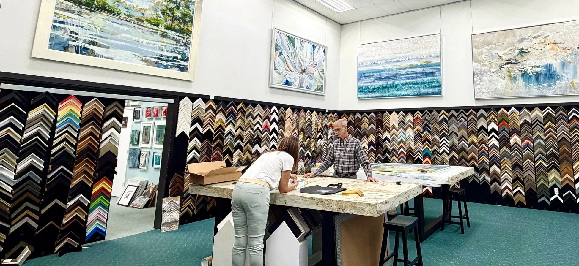 Interior of an art framing store with displays of framed artwork, a wall filled with various frame samples, and two people working at a large work table.