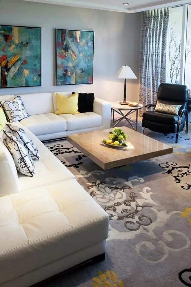 Living room with white sofas, colorful abstract paintings, a black armchair, a side table with a lamp, a coffee table with fruit and flowers, and patterned curtains next to a large window.