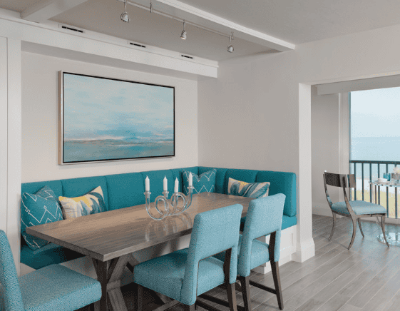 Modern dining room with a long wooden table, blue upholstered chairs, a turquoise banquette with patterned pillows, a candelabra centerpiece, a wall painting of a seascape, and a balcony view in the background.