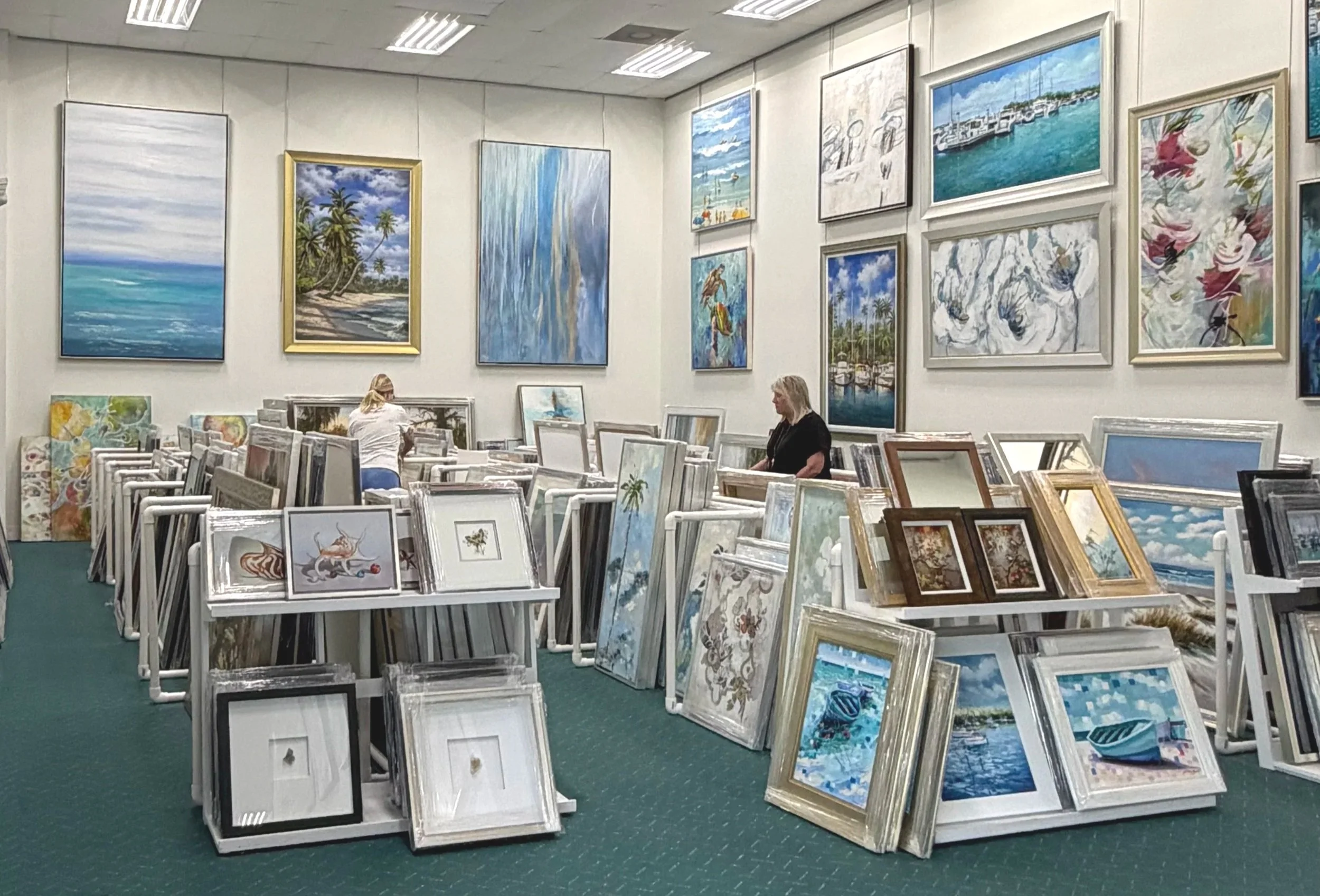An art gallery with paintings on the walls and framed artwork displayed on white tables. The paintings feature beach scenes, ocean views, and tropical landscapes. Two women are browsing the artwork, one with blonde hair and another with gray hair.