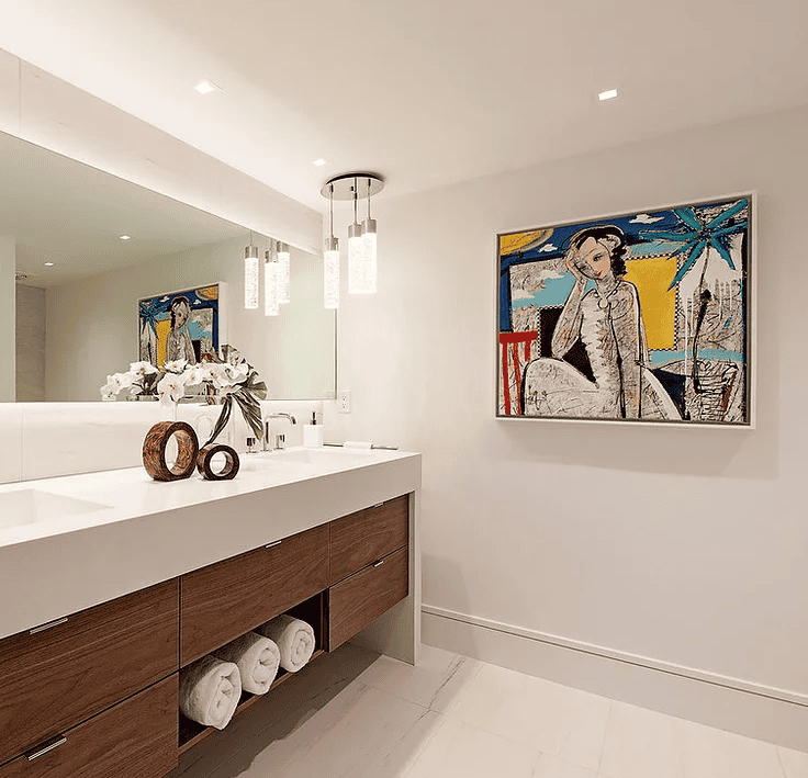 Modern bathroom with white walls and ceiling, a large mirror, a wooden vanity with rolled towels underneath, decorative items on the counter, and colorful artwork of a woman and tropical scenery on the wall.