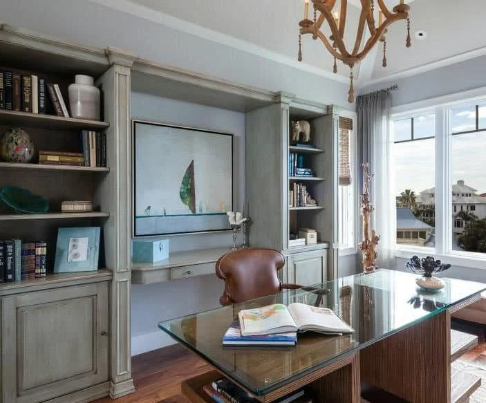 A home office with built-in bookshelves, a glass desk, an open book, a leather chair, a framed painting of a sailboat, and large windows with a view of neighboring houses and palm trees.