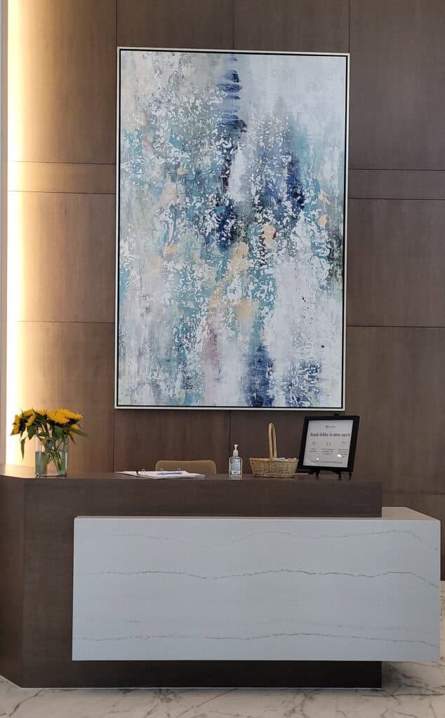 Reception desk with a small bouquet of sunflowers on the left, a bottle of hand sanitizer, a basket, and a sign on the right. Behind the desk is a large abstract painting on a wooden wall.