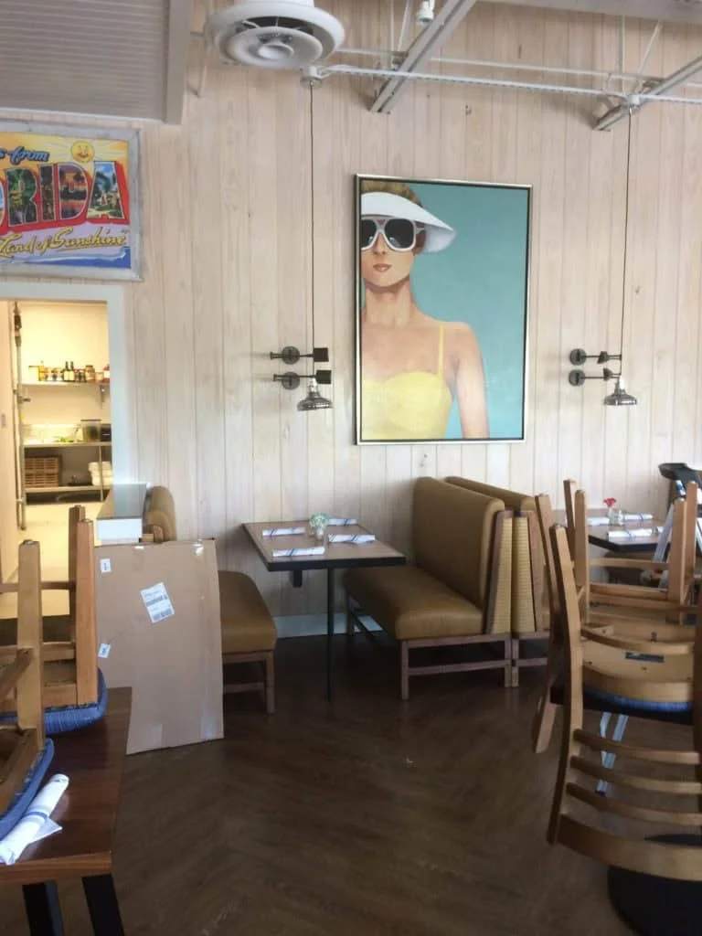 Interior of a restaurant with light-colored wooden walls, a framed painting of a woman wearing sunglasses and a sun hat, and dark wooden flooring. Furniture includes tables, chairs, and booth seating with yellow upholstery. Some chairs are stacked an