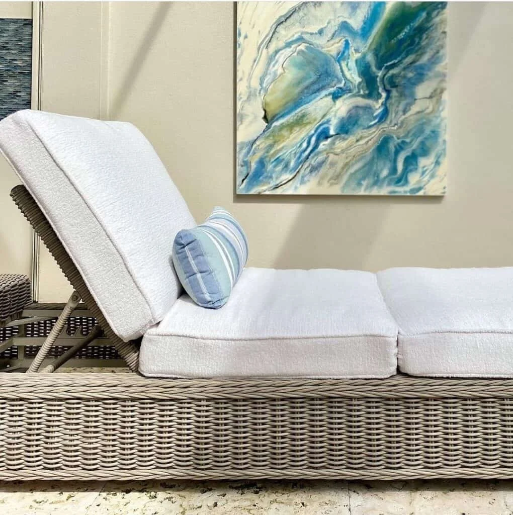 A white lounge chair with a tilted backrest and a blue striped pillow, set in a room with an abstract blue and green painting on the wall behind it.