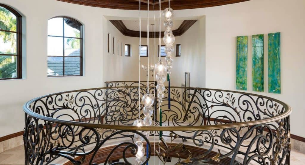 Interior view of a house staircase with ornate wrought iron railing, three large windows letting in natural light, and series of pendant lights hanging from the ceiling.