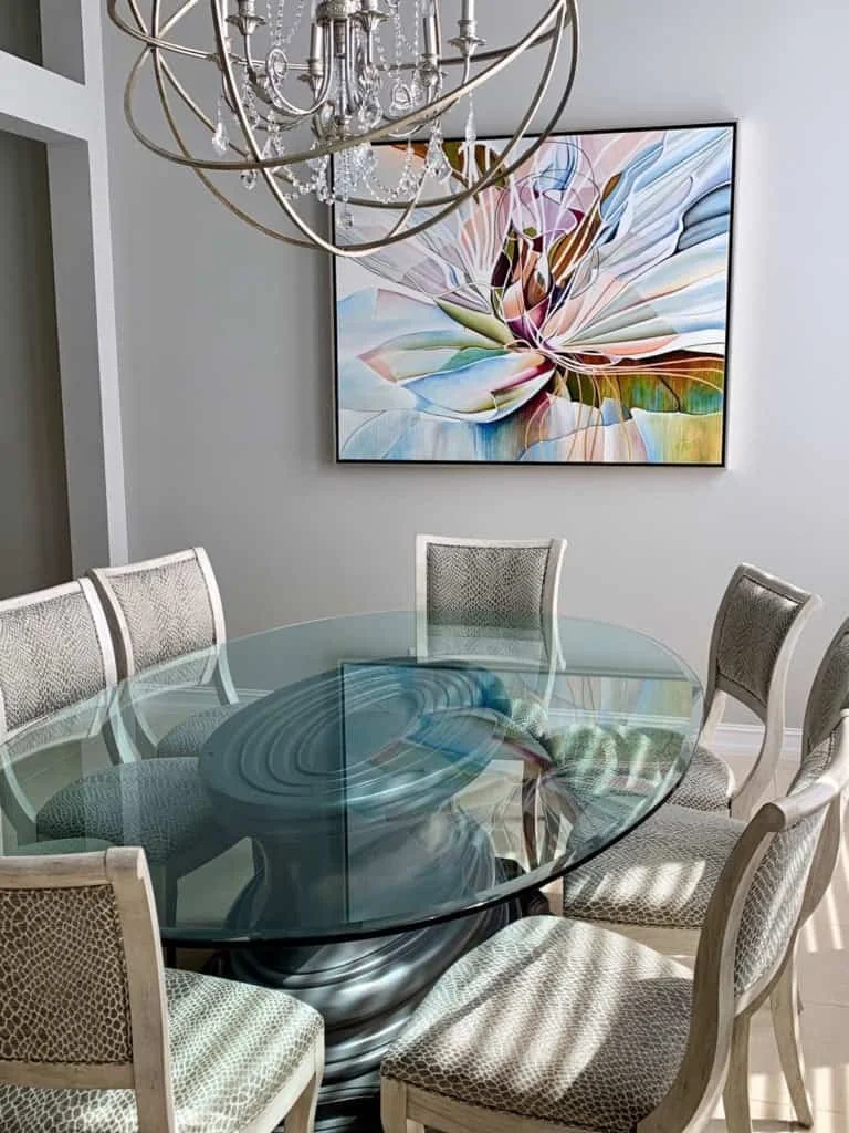 A dining room with a glass-top table, surrounded by six upholstered beige chairs, a modern abstract flower painting on the wall, and a chandelier overhead.