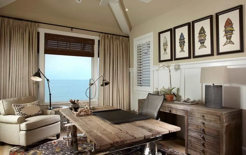 A coastal office with a large window overlooking the ocean, a wooden desk with a black chair, a beige sofa, a wooden sideboard with a lamp, artwork of fishing lures on the wall, and various decor items.