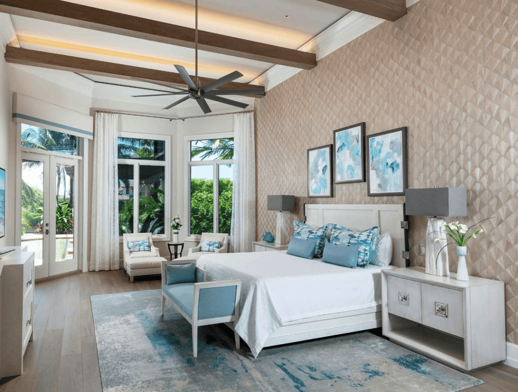 A bright, modern bedroom with a white bed, blue accent pillows, and artwork on the wall. Large windows with white curtains let in natural light. There are two white nightstands with lamps, a blue upholstered bench at the foot of the bed, and a small 