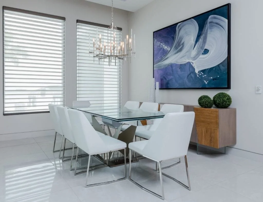 Modern dining room with white chairs around a glass table, a chandelier above, and windows with white blinds. A large abstract painting hangs on the wall, and a wooden sideboard with two decorative green balls on top.