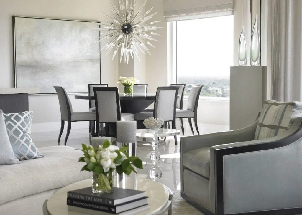 Modern living room with a sitting area and a dining table, decorated with vases and flowers, in a bright space with large windows.