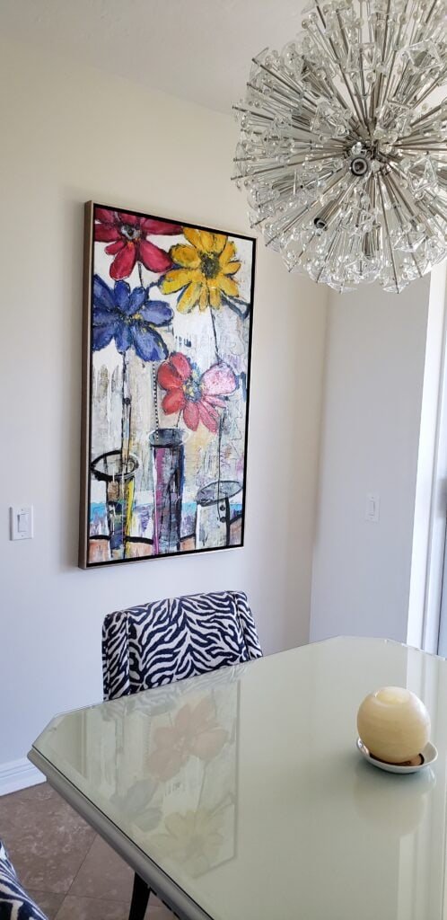 A dining room with a white table, zebra-patterned chairs, a large floral painting on the wall, a modern crystal chandelier overhead, and a round beige decorative object on the table.