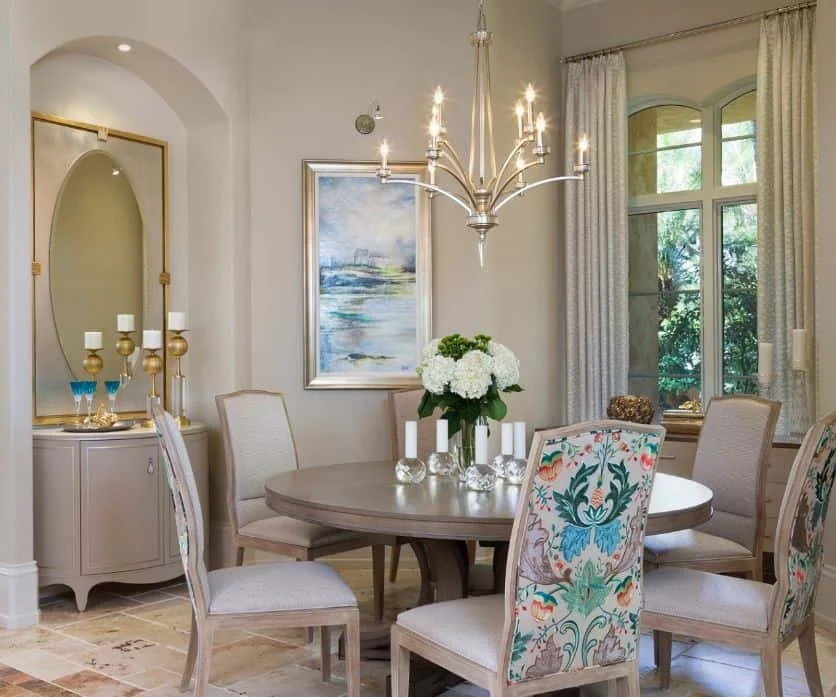 A dining room featuring a round table with a flower centerpiece, surrounded by six upholstered chairs, a chandelier overhead, a side cabinet with decorative items, a framed painting, and a window with curtains letting in natural light.