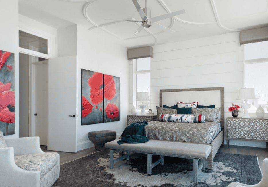 A modern bedroom with white walls, a bed with patterned bedding, colorful pillows, and a long bolster pillow. There are two matching bedside tables with lamps and vases. Artwork with red flowers hangs on the wall, and a bench at the foot of the bed h
