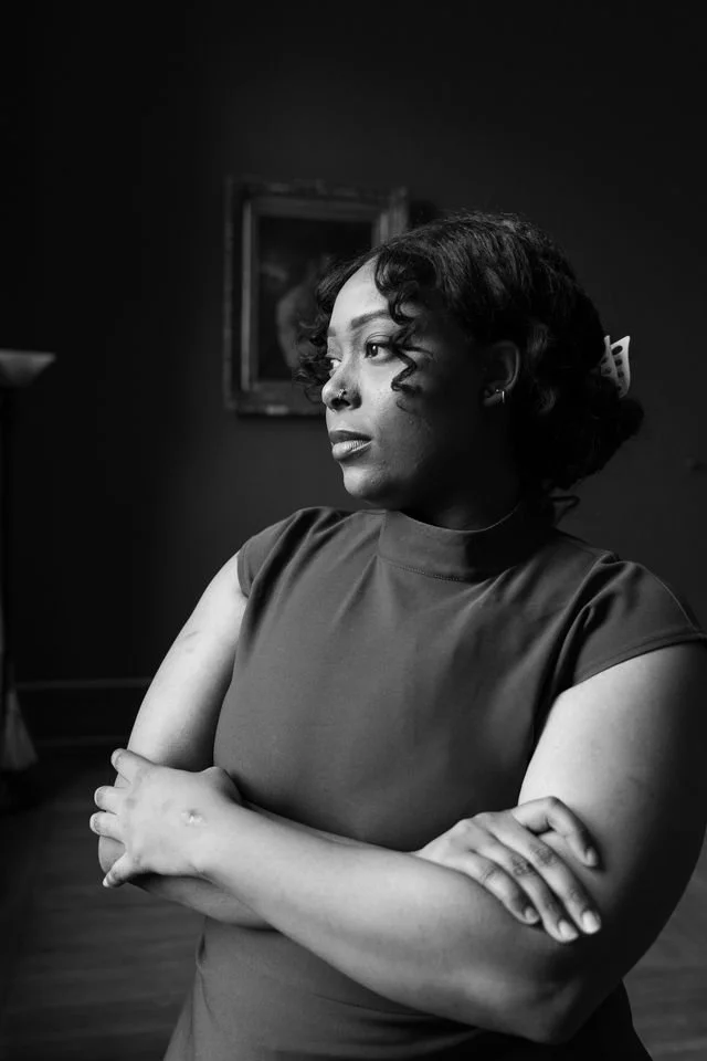 Black and white portrait of a woman with short curly hair, wearing a short-sleeved dress, standing with crossed arms in a room with a blurry painting in the background.