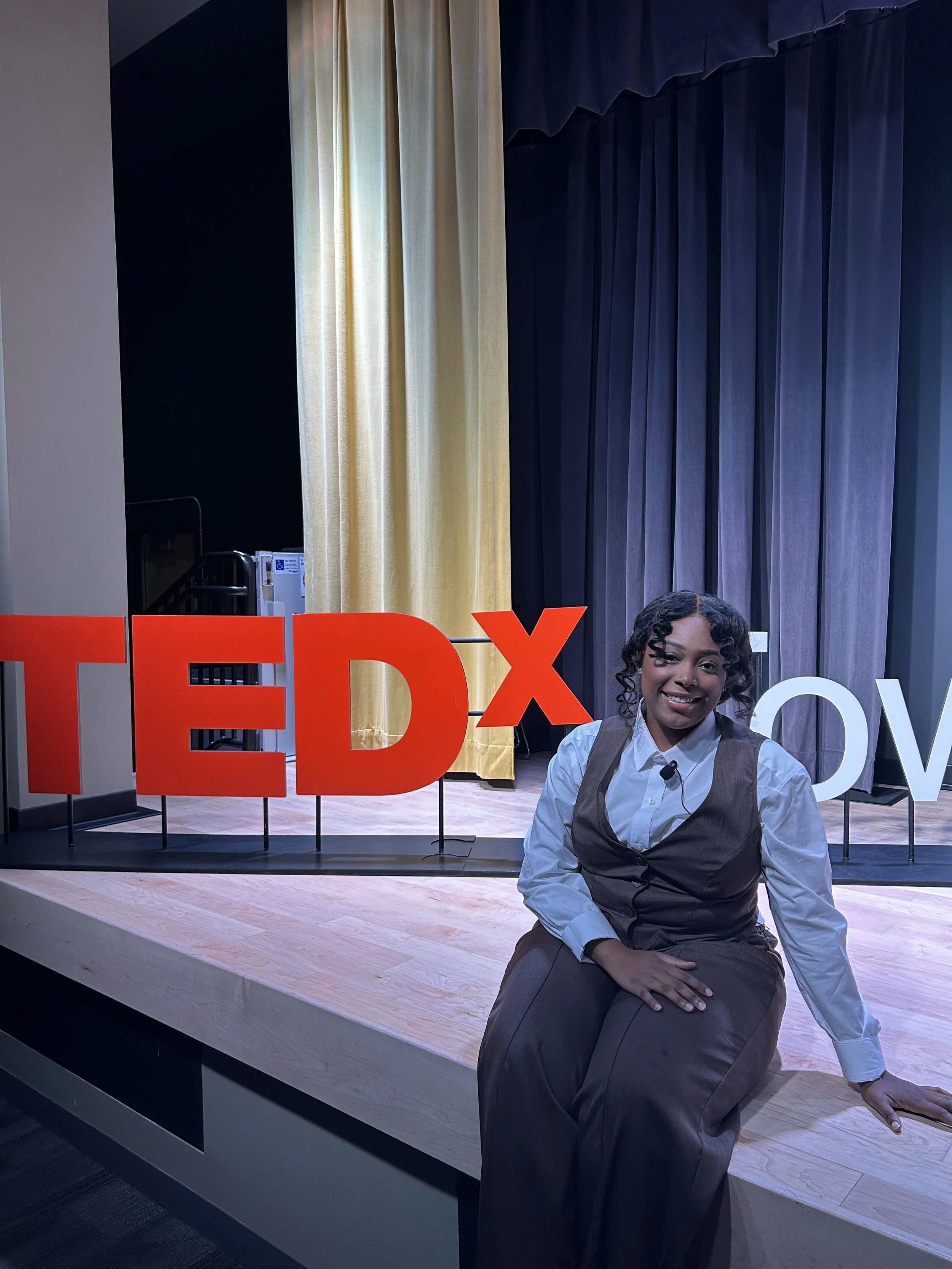 A woman sitting on a stage in front of a large TEDx sign with colored curtains behind her.