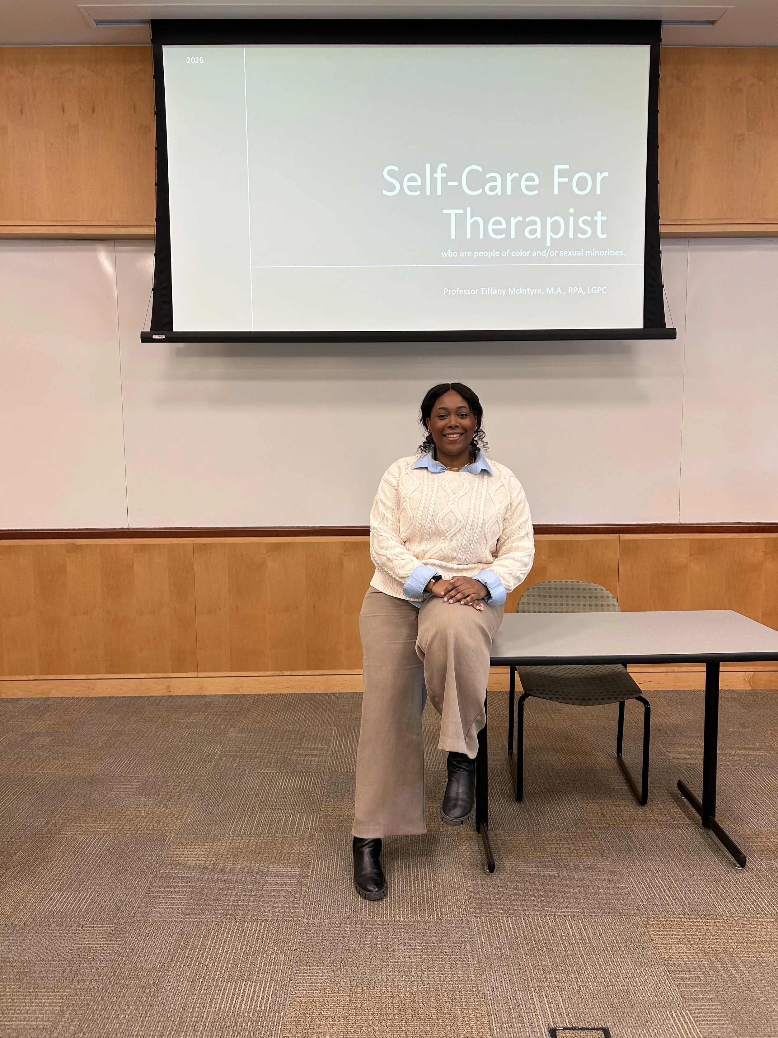 A woman sitting on a raised surface in front of a classroom with a presentation slide on the wall behind her. The slide reads 'Self-Care For Therapist' by Professor Tiffany McIntyre.