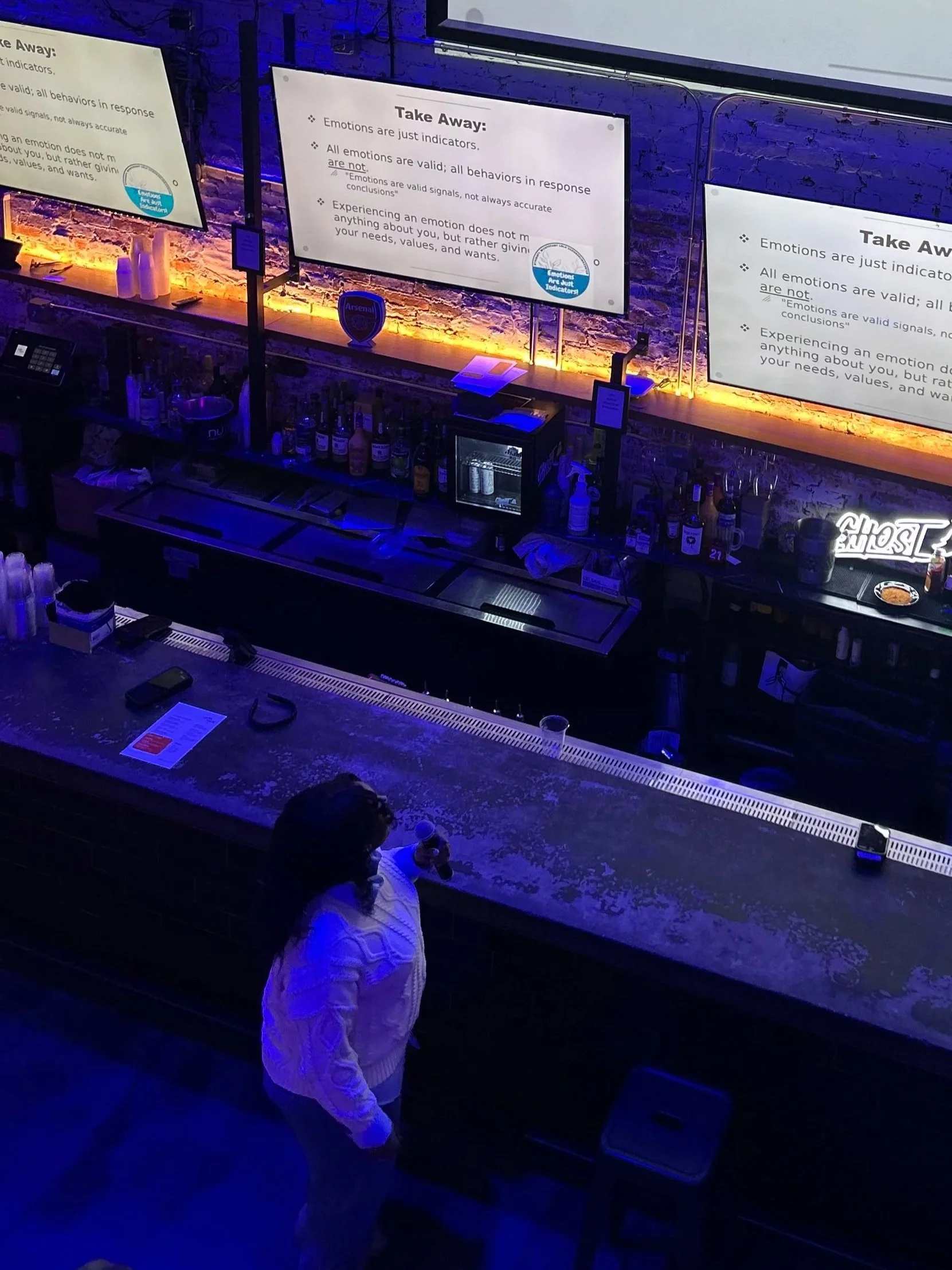 A person with dark hair wearing a white jacket standing at a bar, drinking from a glass, near a person with curly hair wearing a light-colored sweater. The bar has bottles of alcohol and two large screens displaying text about emotions and response indicators. The lighting is dim with blue and orange hues.