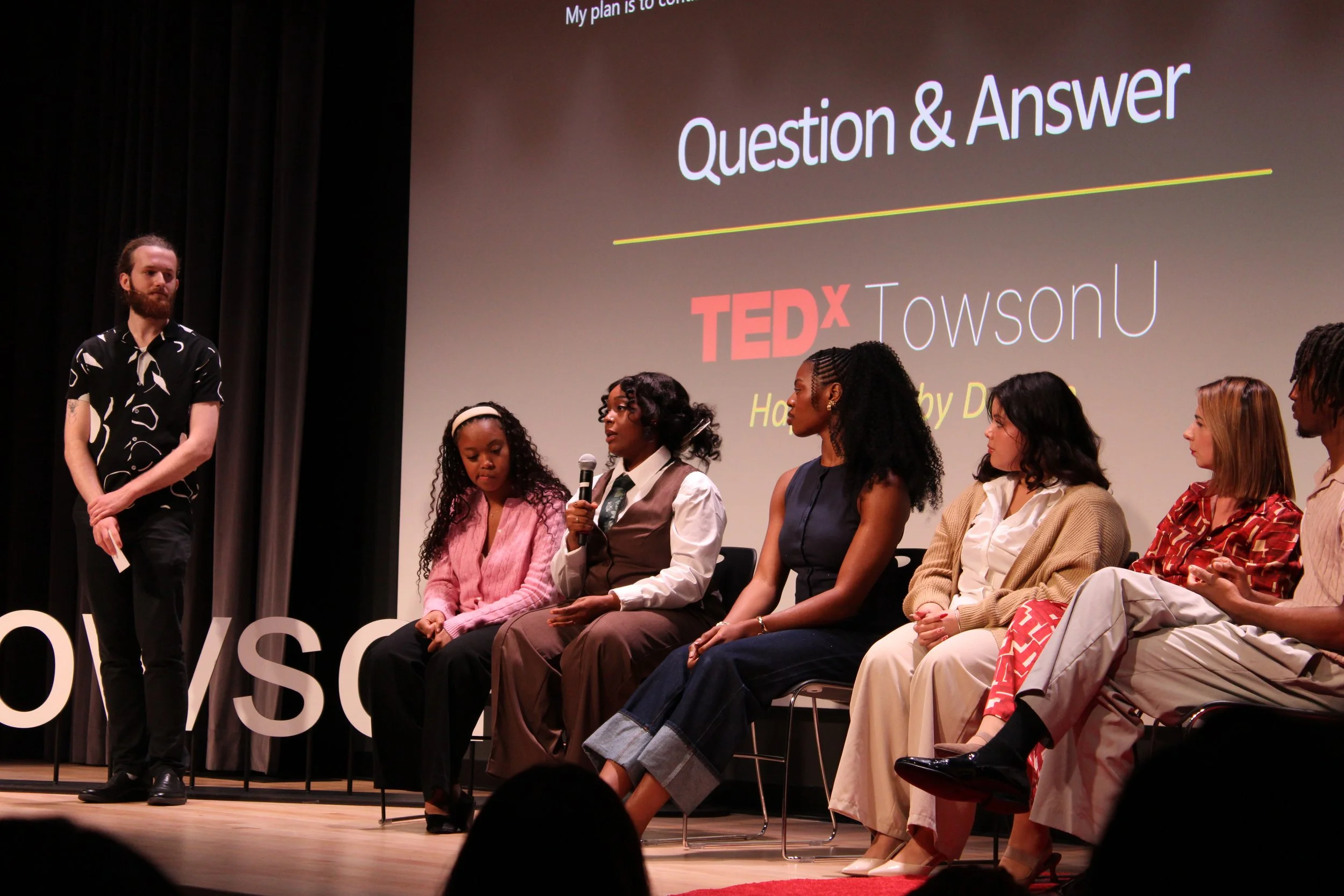 A panel discussion at a TEDx event with six women seated on stage, one holding a microphone, and a young man standing next to them. The background displays a large screen with the words 'Question & Answer' and the TEDxTowsonU logo.