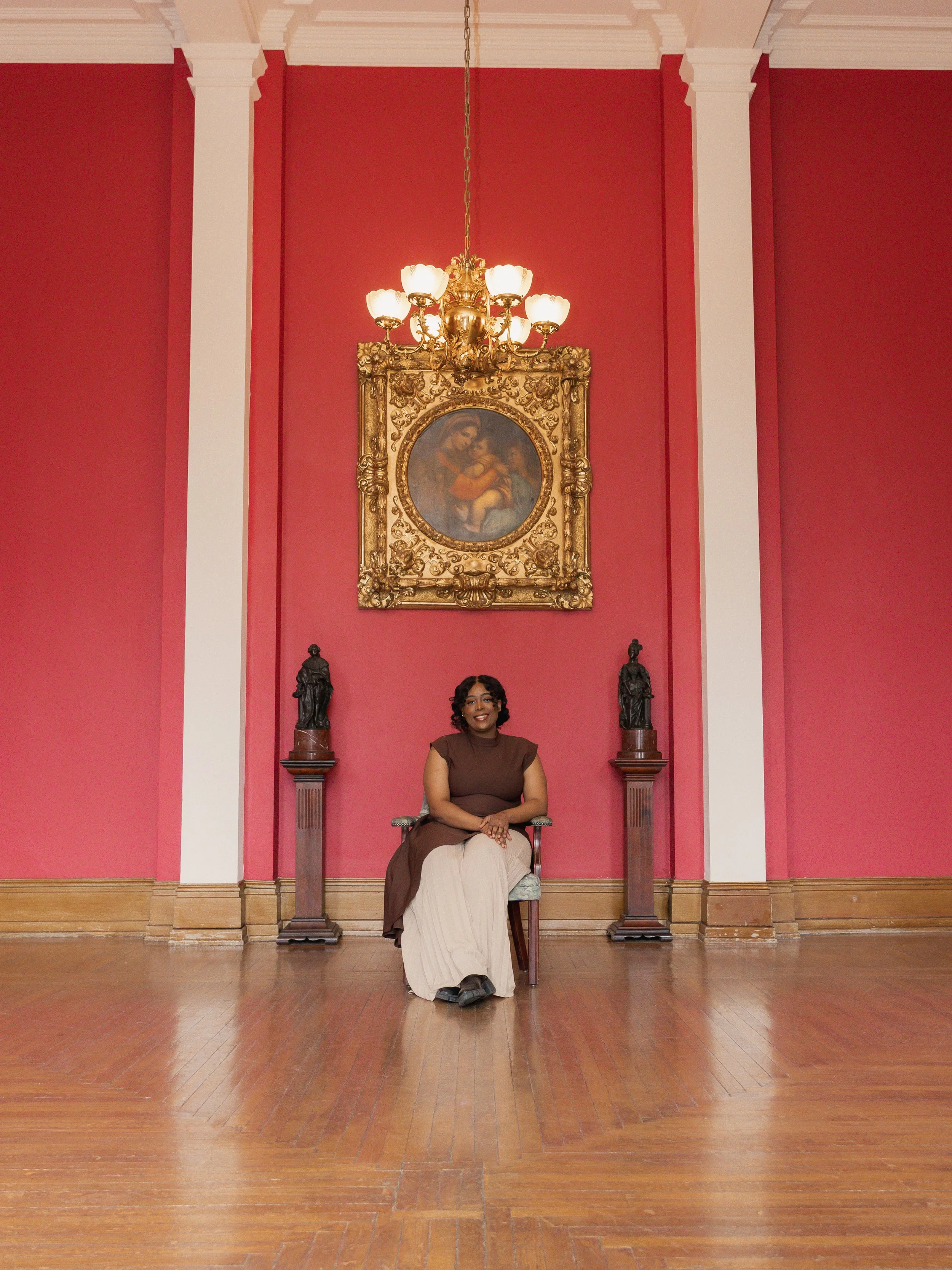 A woman sits on a chair with a detailed armrest in a room with pink walls, white trim, and wooden flooring. Behind her, an ornate gold-framed painting and two statues are displayed, with a chandelier hanging from the ceiling.