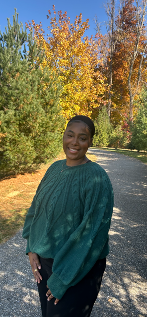 A woman smiling outdoors on a paved trail with fall-colored trees in the background.