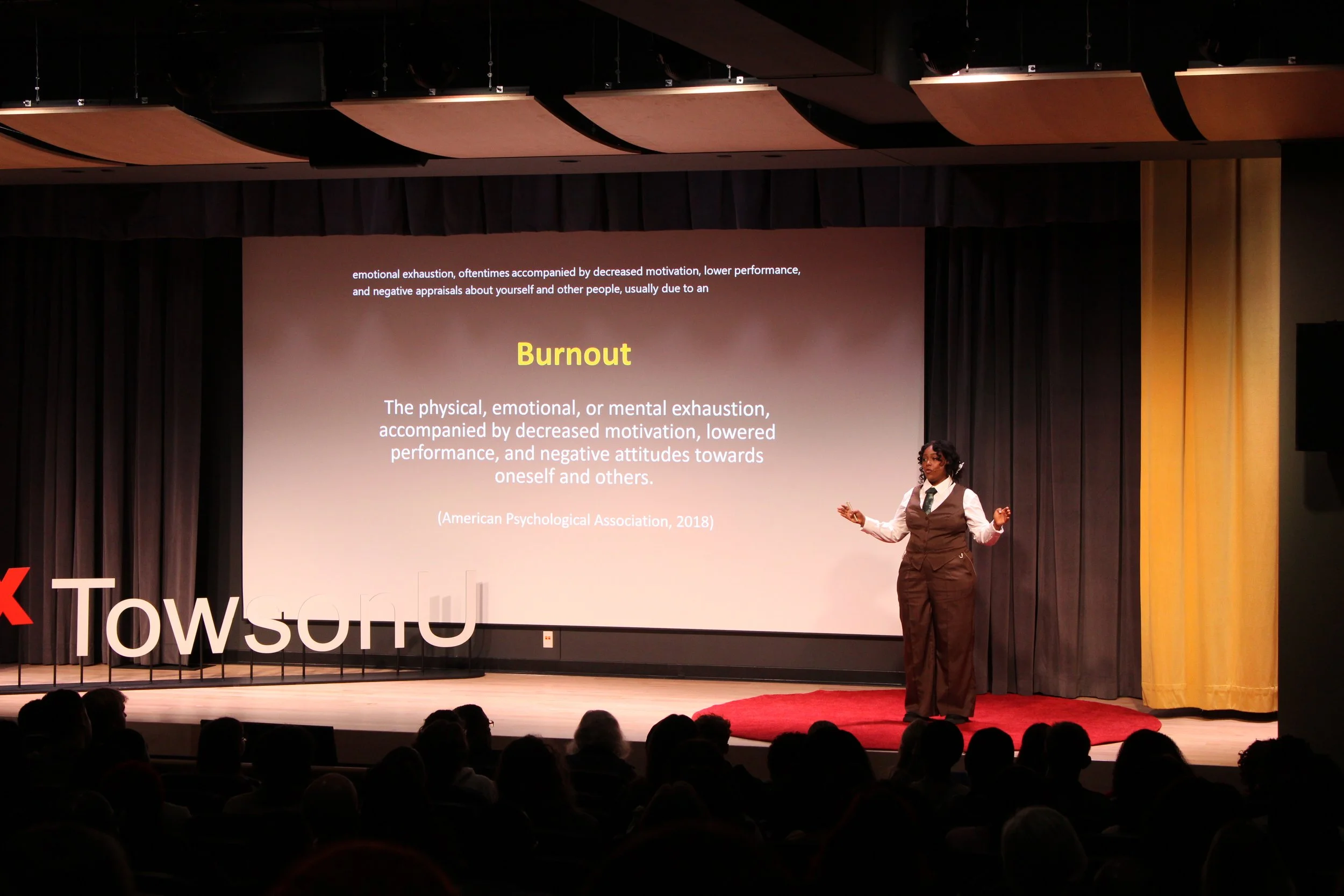 A woman in formal attire giving a presentation on a stage with a large screen behind her displaying information about burnout, in a theater or conference hall filled with an audience.