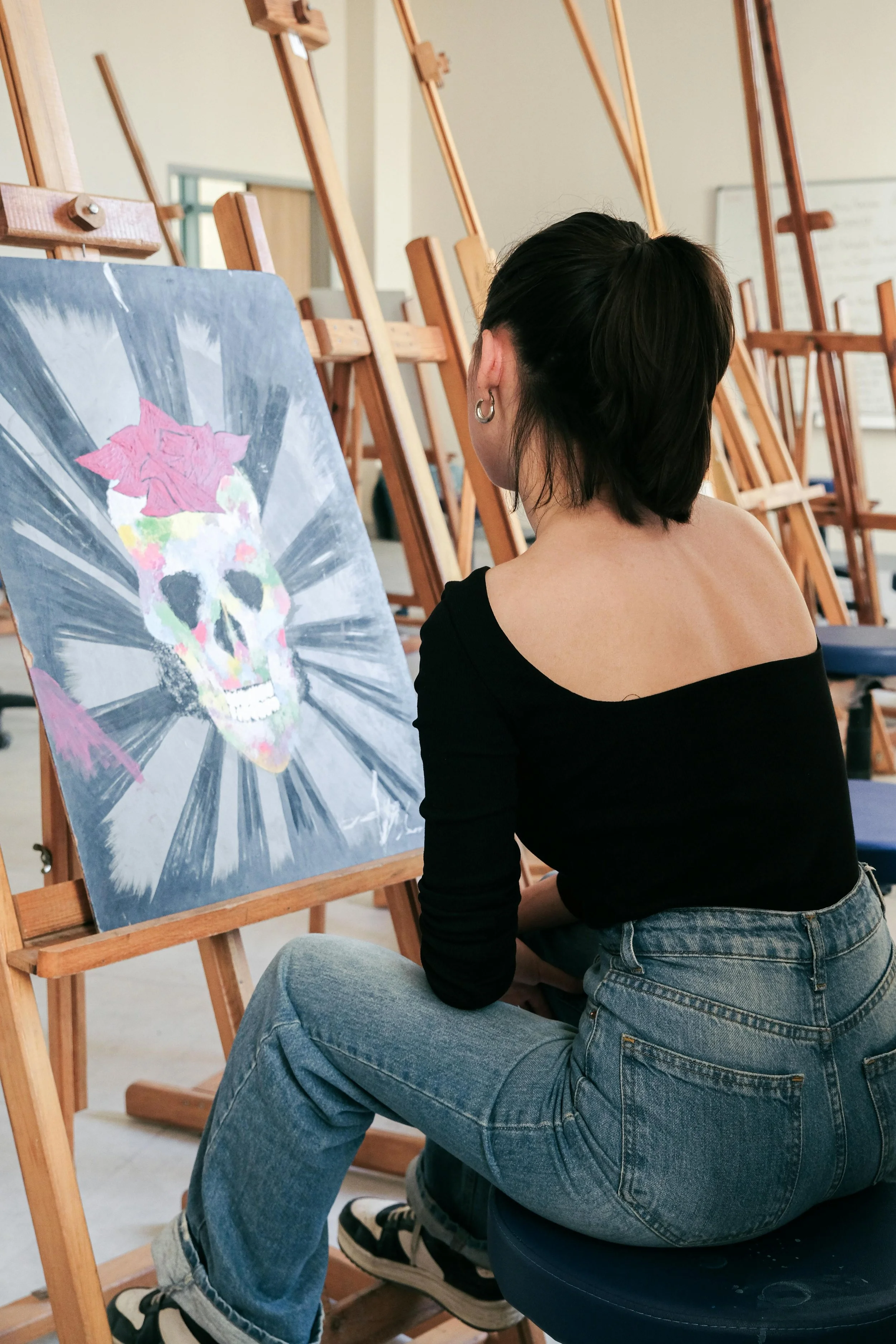 A person with short dark hair, wearing a black off-shoulder top and blue jeans, sitting on a black stool in front of an easel, observing a colorful skull painting with a pink flower on its head, in an art studio with blank easels in the background.
