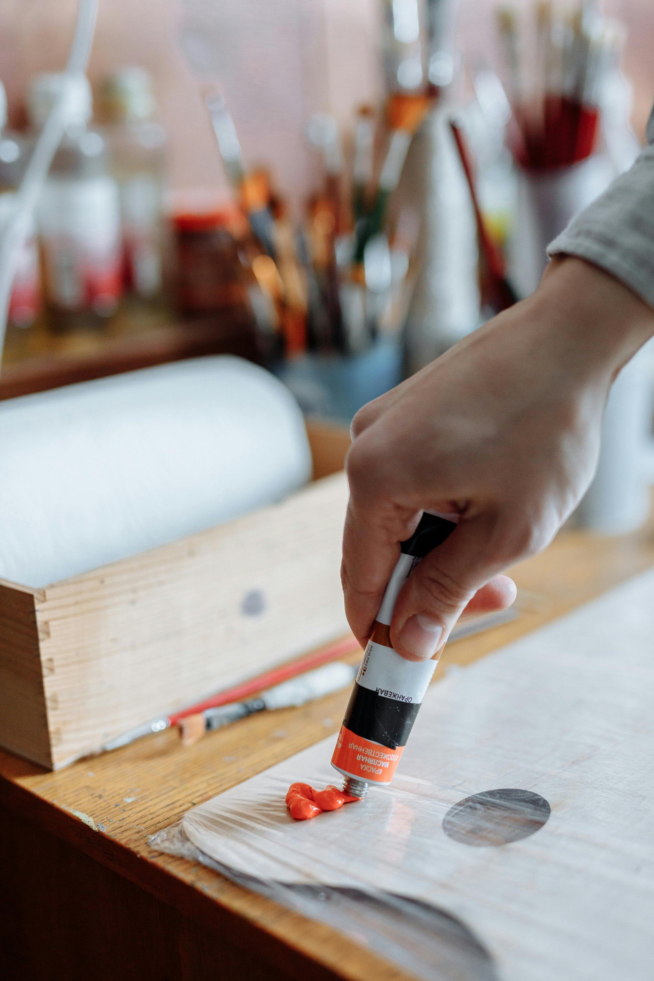 A person is squeezing orange-colored paint from a tube onto a piece of wood, with various art supplies blurred in the background.