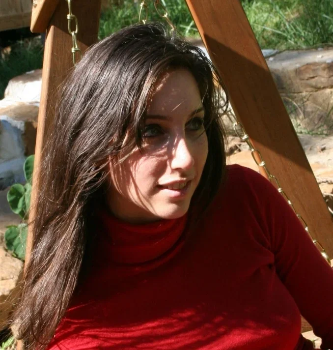 A woman with long dark hair wearing a red turtleneck top sitting outdoors near a wooden structure with chains, sunlight casting shadows on her face.
