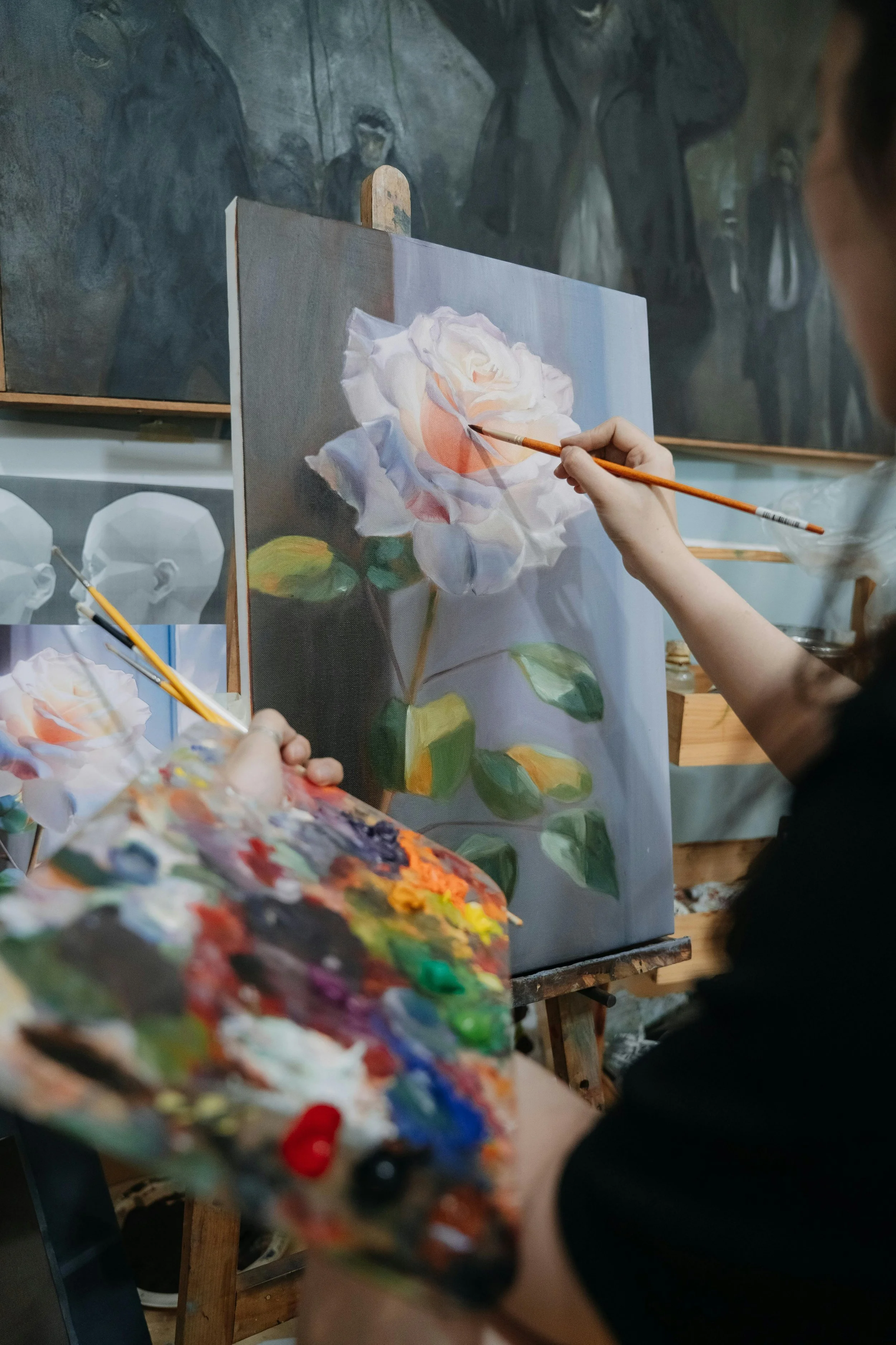 A person painting a realistic white rose with green leaves on a canvas, with a palette of various paint colors and brushes, in an art studio surrounded by other artworks.