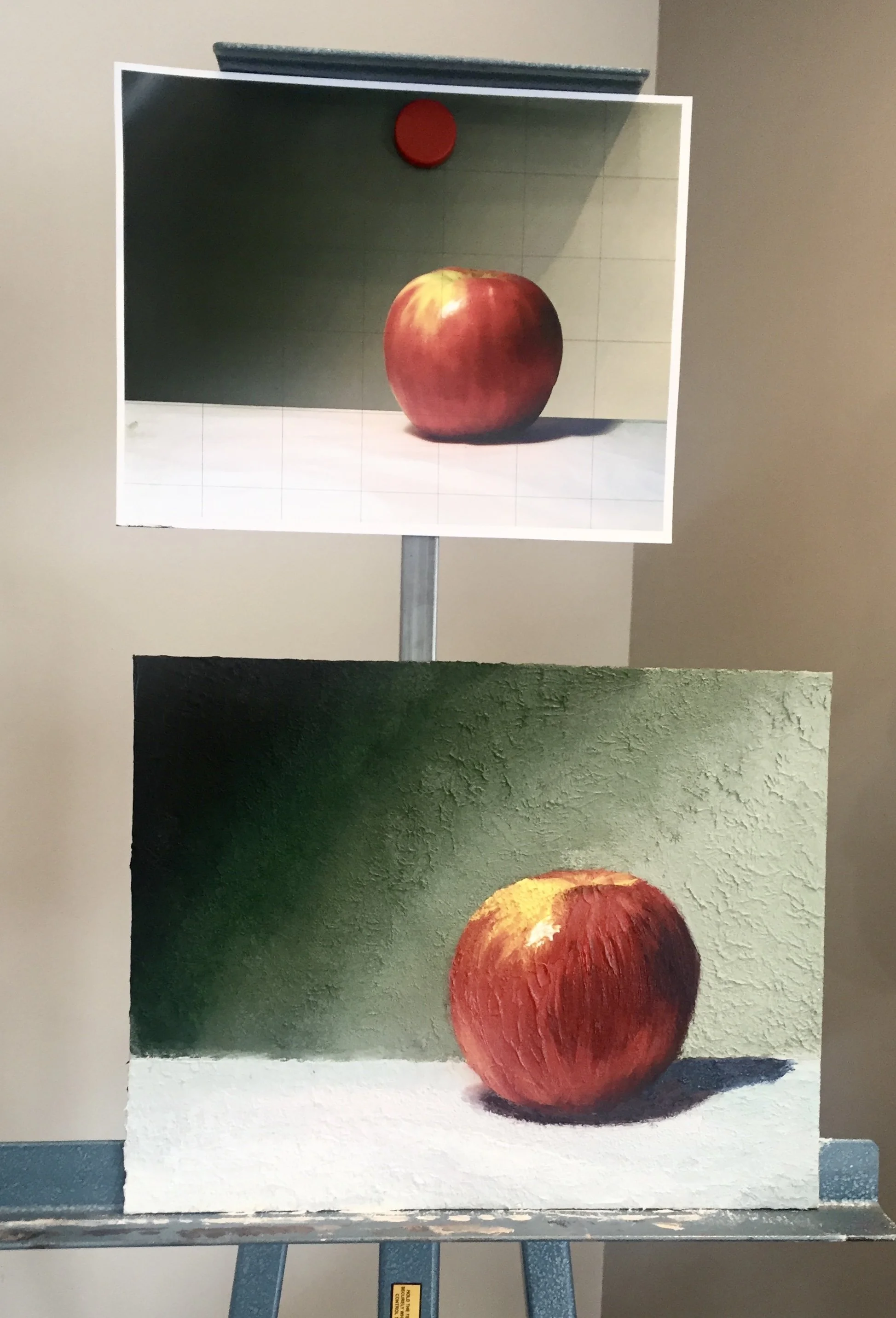 Photograph of a realistic painting of a red apple with yellowish streaks, on a white surface, with a plain background.