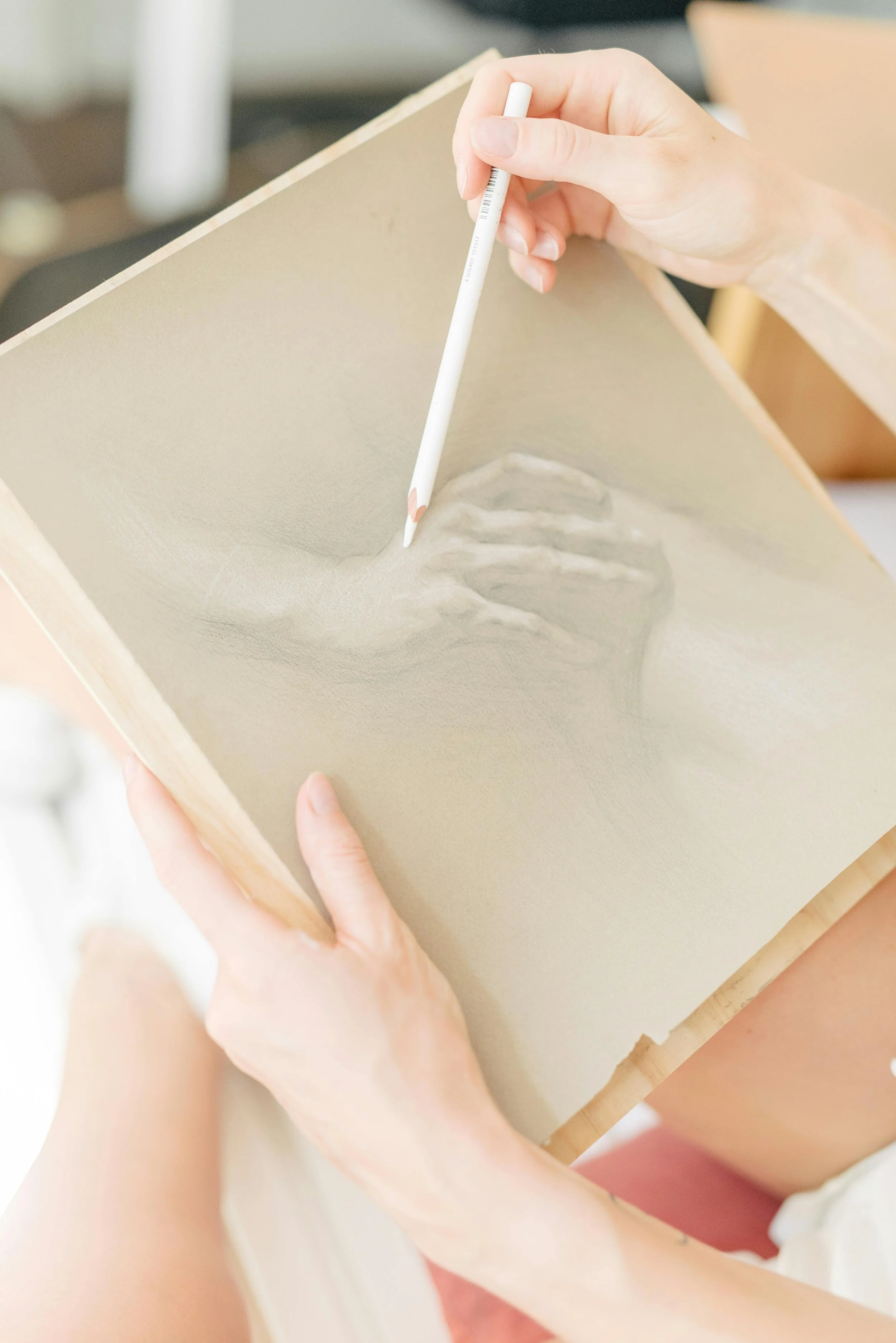 Close-up of a person using a white colored pencil to draw or shade on a textured beige-colored surface, with hands holding the surface and pencil. The background is blurred.