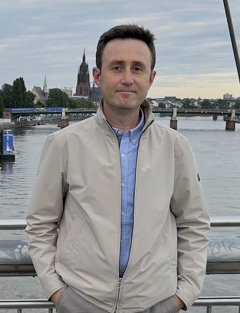 A man standing on a bridge with a river and cityscape in the background, including a church steeple and bridge over the river, under a cloudy sky.