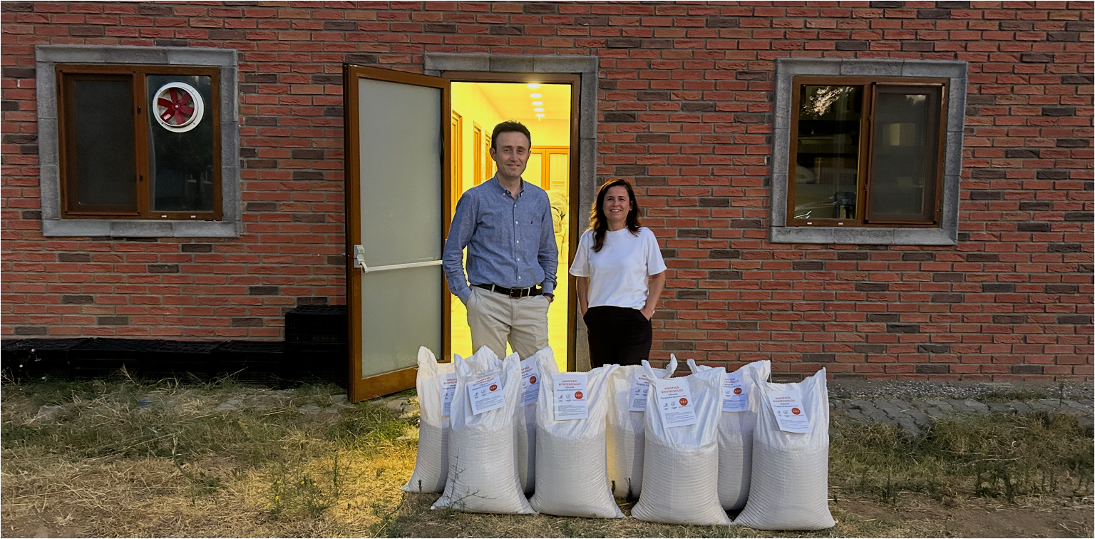 A man and a woman stand outside a brick building next to bags of what appears to be grain or flour. The man is wearing a light blue shirt and khakis, and the woman is wearing a white top and dark skirt. The bags are lined up on the ground in front of them, with labels attached.
