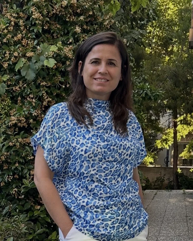 Woman with shoulder-length brown hair wearing a blue animal print top, standing outdoors in front of green foliage and flowering bushes.