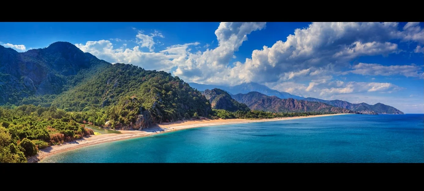 Panoramic view of a rocky island with green mountains, sandy beach, and blue water, under a partly cloudy sky.