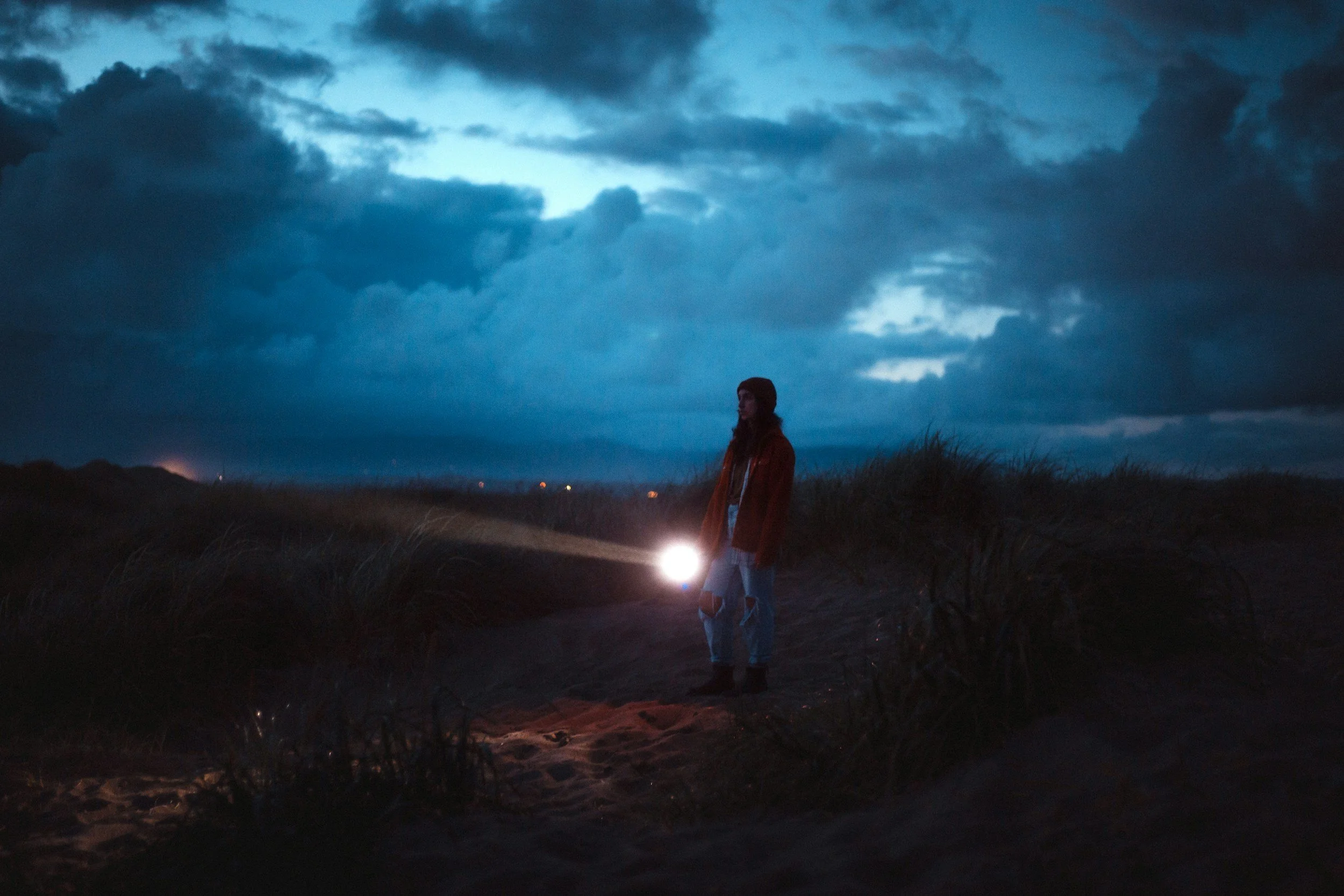 A woman standing on a sandy path outdoors at dusk, holding a flashlight, with dark clouds overhead and a distant horizon with lights.