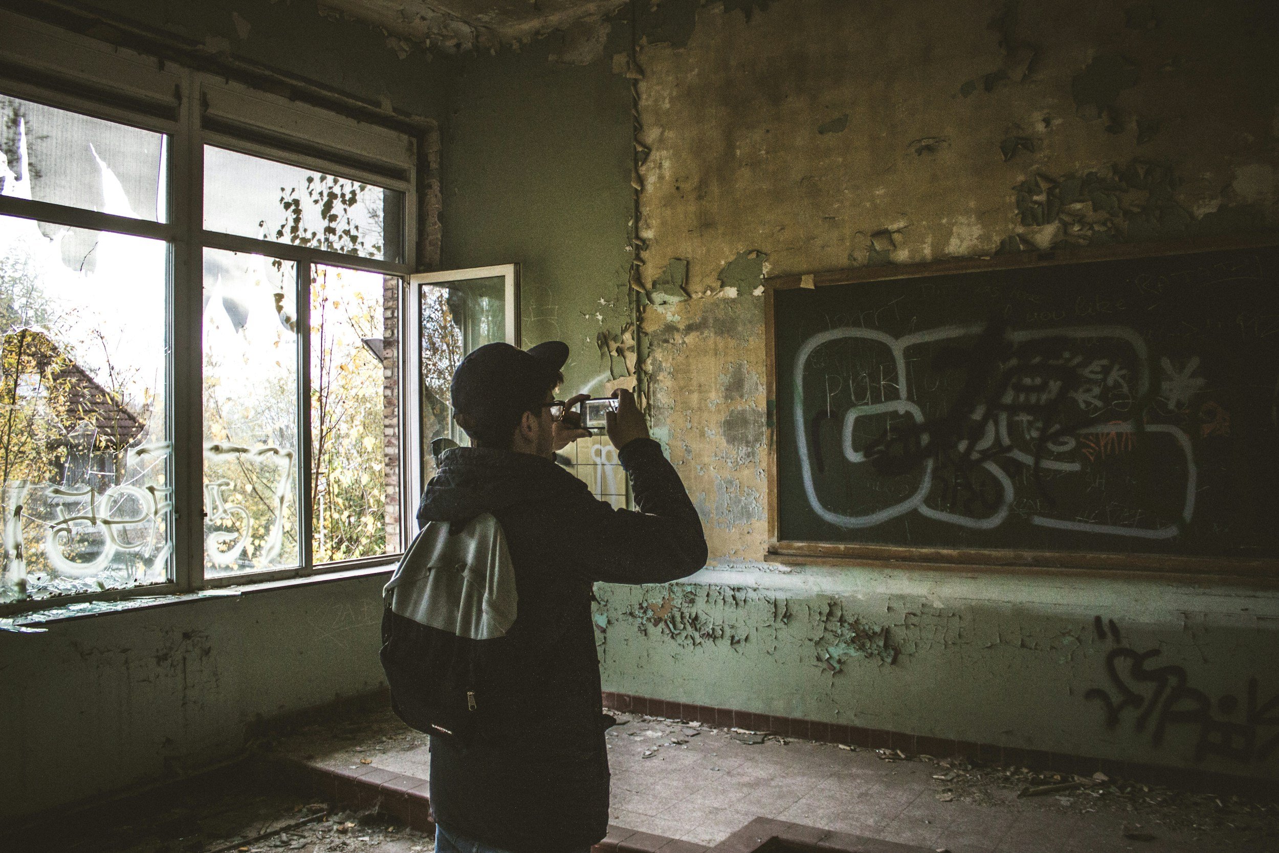 Person taking a photo inside a dilapidated room with peeling paint, broken window, and graffiti on a chalkboard.