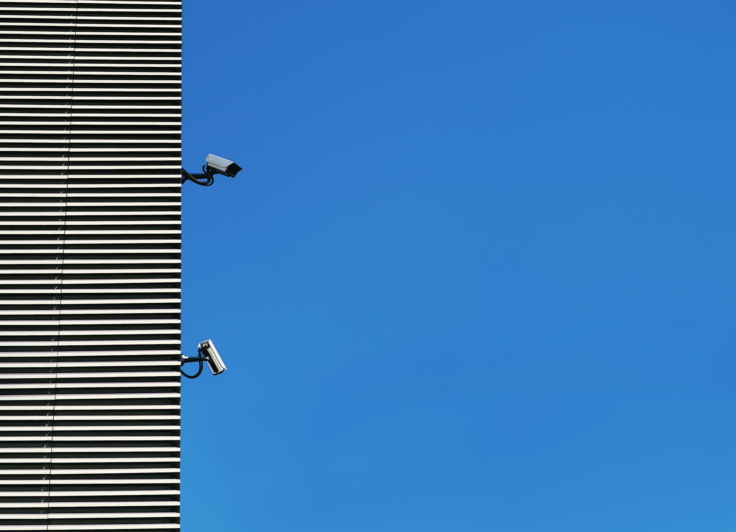 A portion of a building with horizontal silver and black striped panels on the left side of the image. Two security cameras are mounted on the building, extending outward, with the background being a clear blue sky.